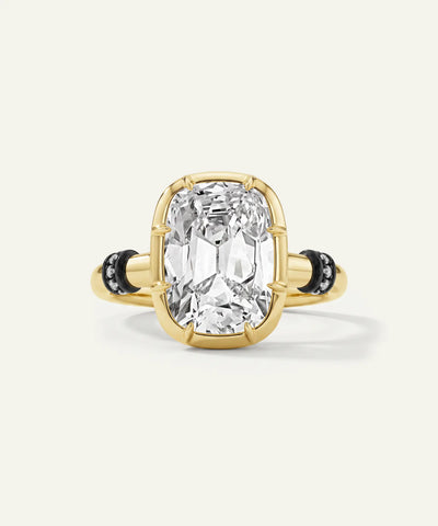 A yellow gold ring featuring a large cushion-cut clear gemstone in a prong setting, accented by small black stones on the band, set against a light cream background.