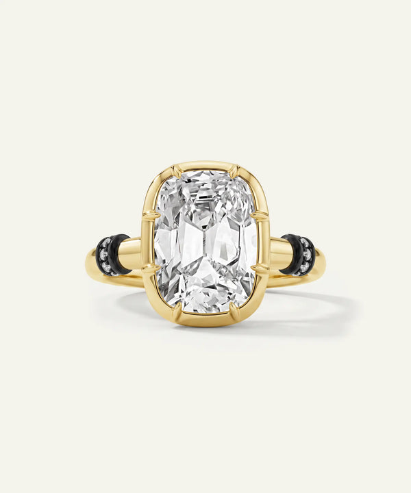 A yellow gold ring featuring a large cushion-cut clear gemstone in a prong setting, accented by small black stones on the band, set against a light cream background.