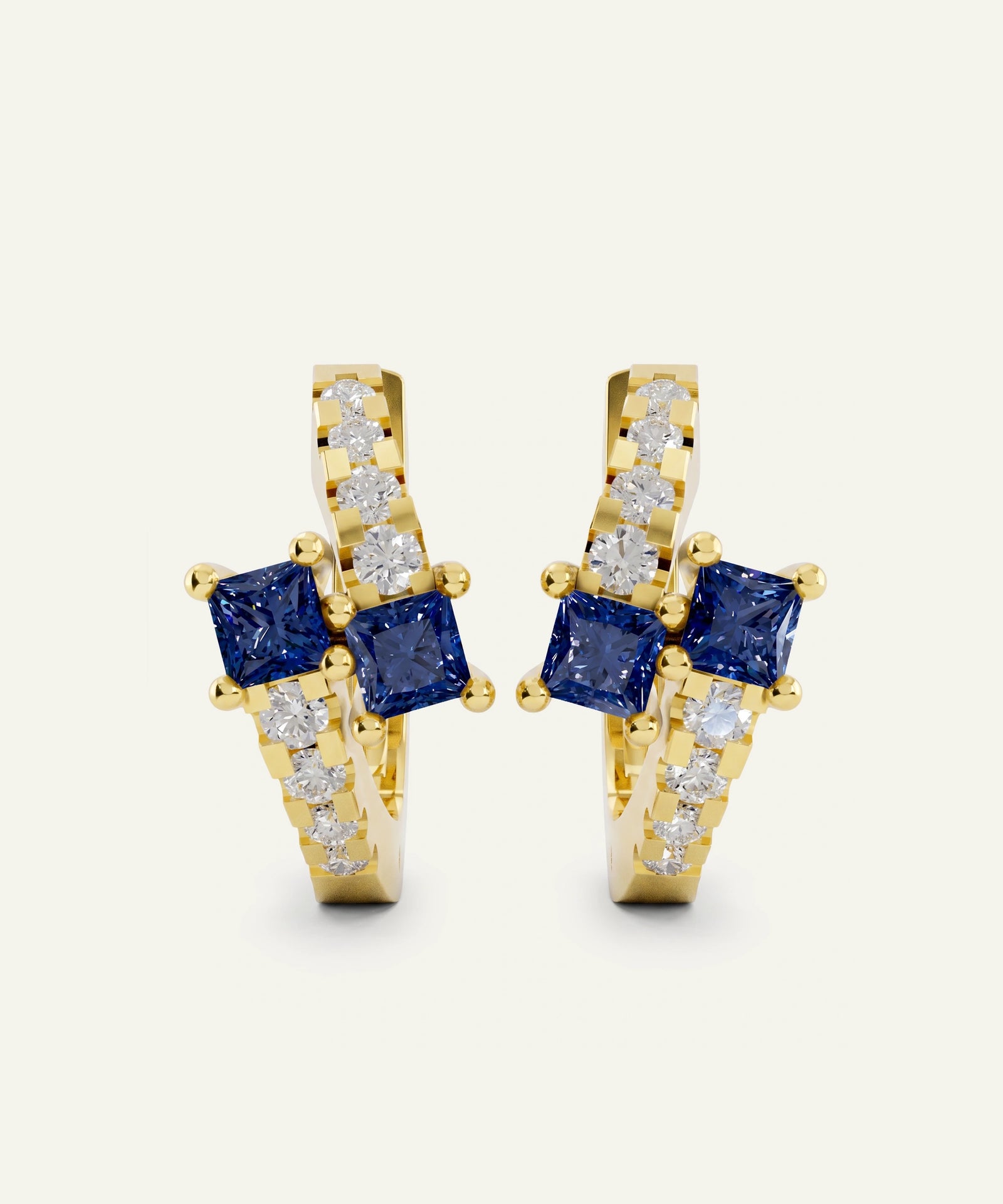 A pair of gold huggie hoop earrings featuring square blue sapphires and pavé-set clear stones, shown on a light cream background.