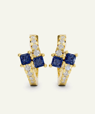 A pair of gold huggie hoop earrings featuring square blue sapphires and pavé-set clear stones, shown on a light cream background.
