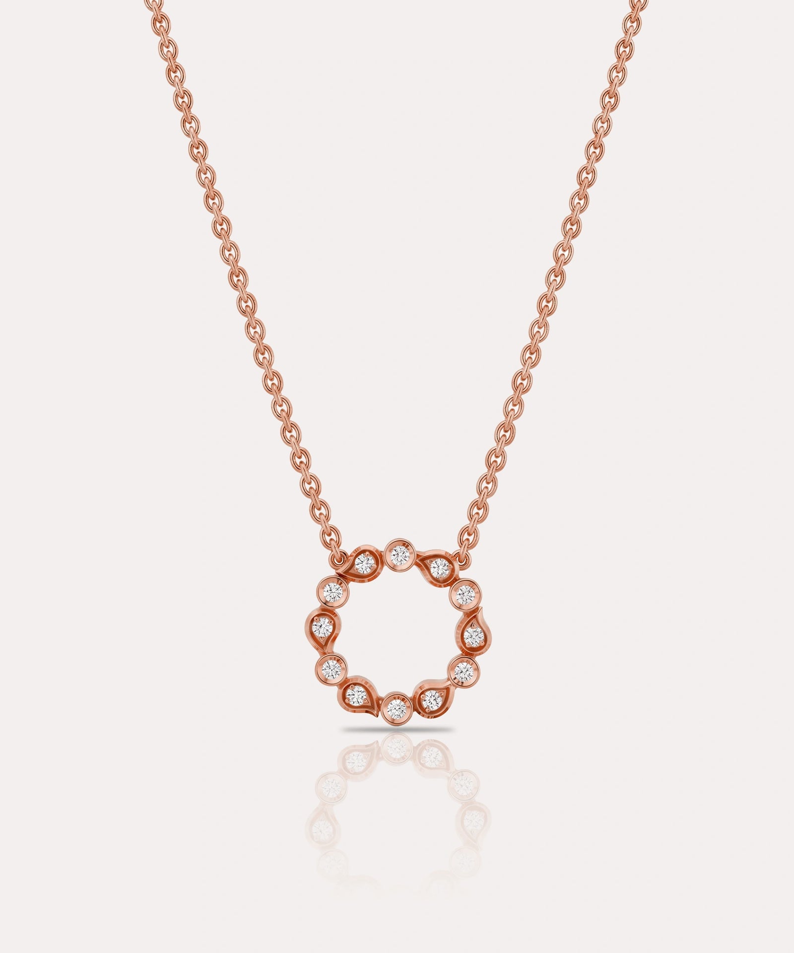 A delicate rose gold chain necklace featuring a circular openwork pendant adorned with small clear gemstones, set against a light off-white background with a soft reflection below.