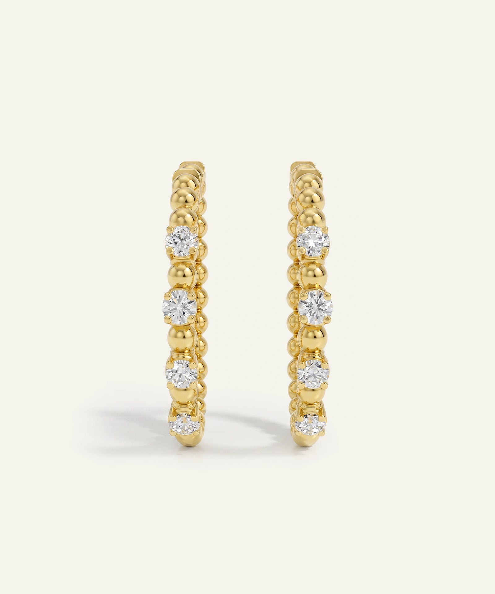 A pair of gold huggie hoop earrings with a textured beaded band and a row of clear round-cut gemstones, shown on a light cream background.
