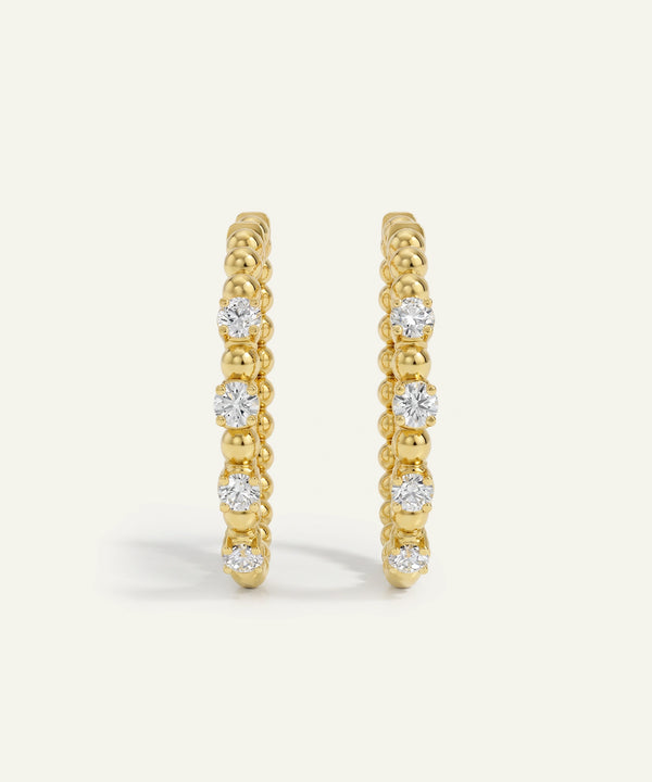 A pair of gold huggie hoop earrings with a textured beaded band and a row of clear round-cut gemstones, shown on a light cream background.