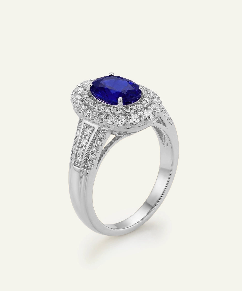 A silver halo ring featuring a round-cut deep blue sapphire center stone surrounded by a halo of small clear stones, with a polished silver band decorated with pavé-set diamonds, shown on a light cream background.