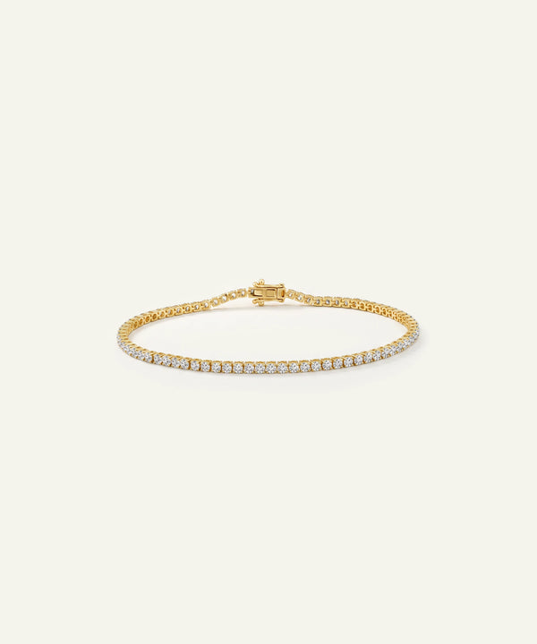 A delicate gold tennis bracelet with a single row of small clear stones, displayed on a light cream background.