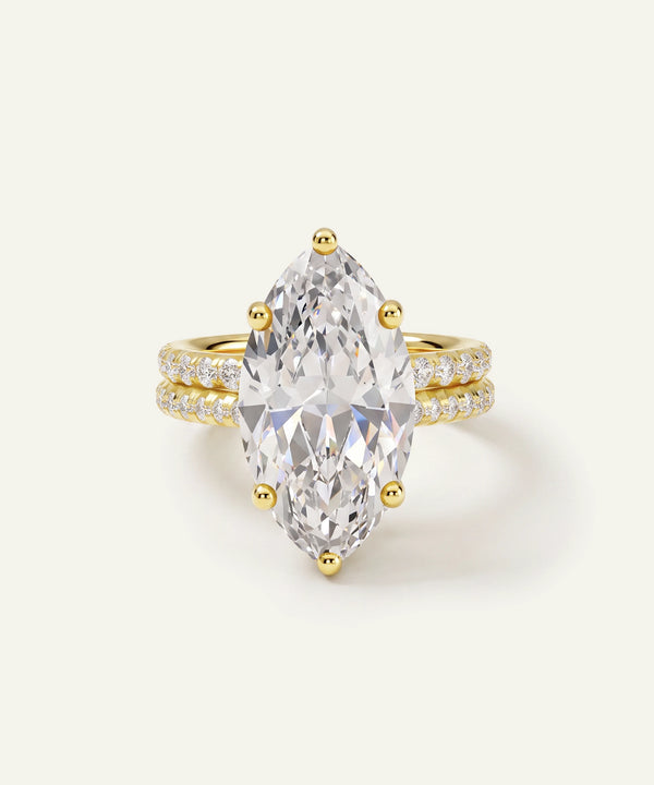 A yellow gold ring featuring a large marquise-cut clear gemstone in a prong setting, with a double-row pavé band accented by small round diamonds, shown on a light cream background.