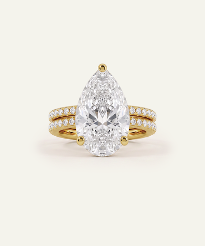 A pear-cut diamond engagement ring with a yellow gold band accented by pavé diamonds, shown on a light cream background.