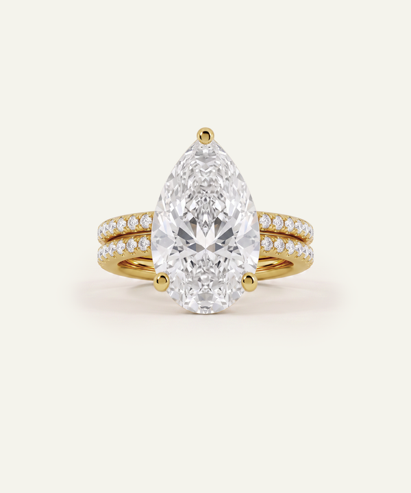 A pear-cut diamond engagement ring with a yellow gold band accented by pavé diamonds, shown on a light cream background.
