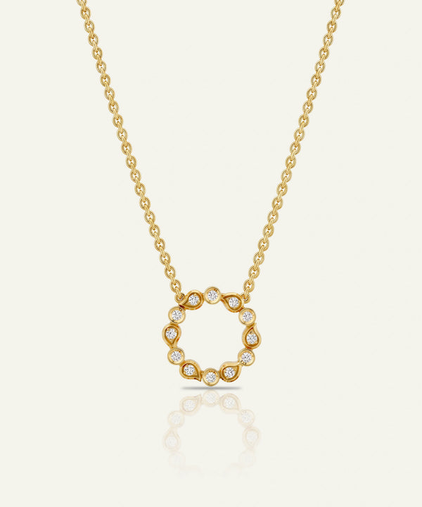 A delicate gold chain necklace featuring a circular pendant with small clear stones set along the edge, displayed against a light cream background with a soft reflection below.