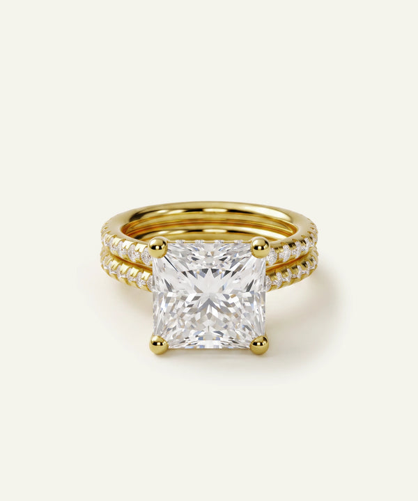 A yellow gold ring set featuring a large princess-cut diamond center stone with a pave diamond band, photographed on a clean off-white background.