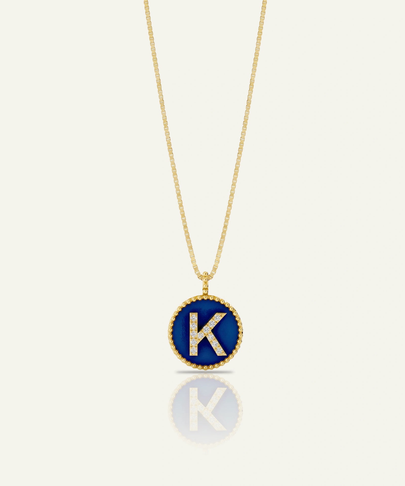 A gold chain necklace with a circular pendant featuring a deep blue enamel background, a gold beaded border, and a raised gold letter 'K' accented with small clear stones, shown on a light cream background with a soft reflection below.