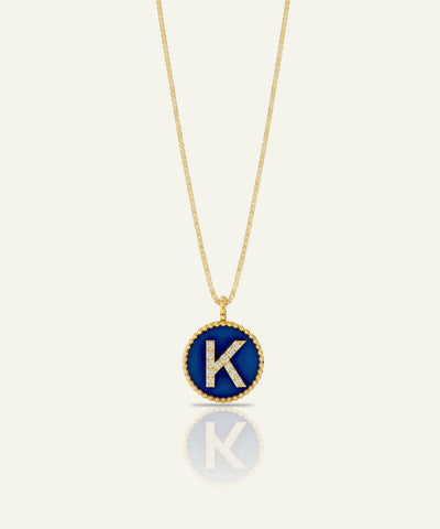 A gold chain necklace with a circular pendant featuring a deep blue enamel background, a gold beaded border, and a raised gold letter 'K' accented with small clear stones, shown on a light cream background with a soft reflection below.