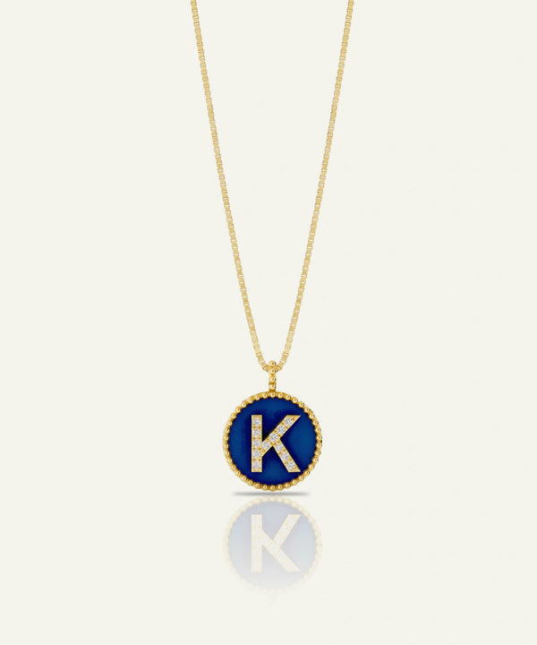 A gold chain necklace with a circular pendant featuring a deep blue enamel background, a gold beaded border, and a raised gold letter 'K' accented with small clear stones, shown on a light cream background with a soft reflection below.
