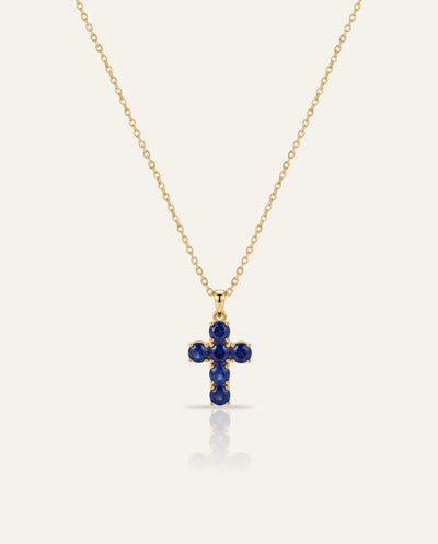 A delicate gold chain necklace with a small cross pendant set with deep blue gemstones, displayed against a light cream background with a soft reflection below.