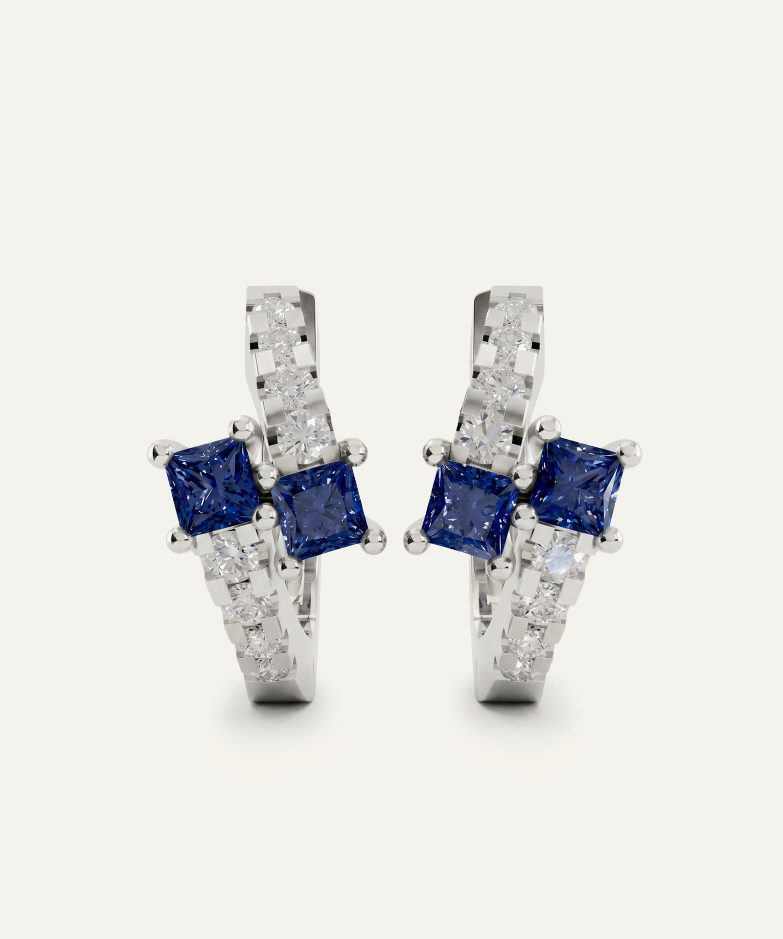 A pair of silver-toned huggie hoop earrings featuring two princess-cut blue sapphires and pavé-set clear stones, shown on a light off-white background.