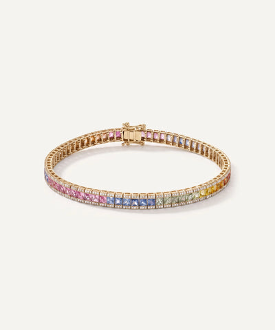 A gold tennis bracelet featuring a row of colorful pave gemstones in pink, blue, yellow, and orange, displayed on a clean off-white background.