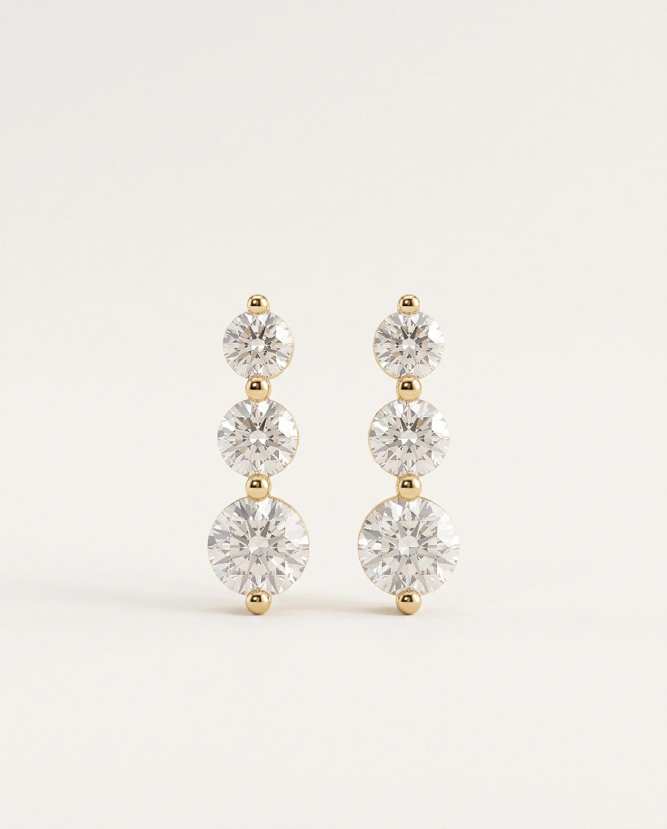 A pair of gold drop earrings featuring three round clear gemstones in each earring, set against a plain off-white background.