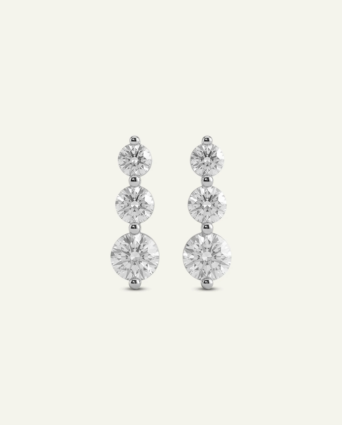 A pair of silver-toned drop earrings featuring three round-cut clear gemstones in each earring, displayed against a solid off-white background.