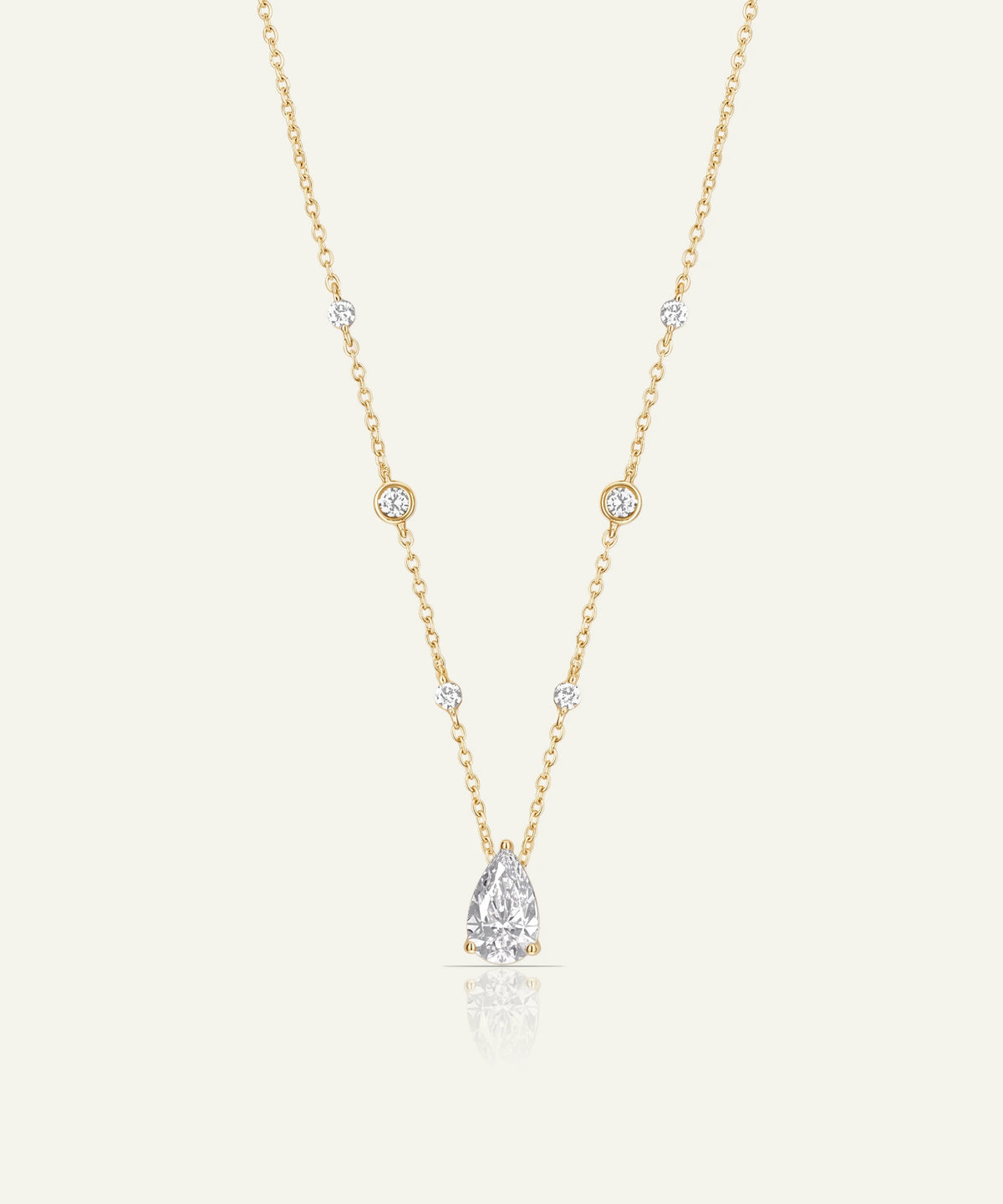 A delicate yellow gold necklace featuring a pear-cut diamond pendant and small round diamond accents along the chain, displayed against a light cream background.