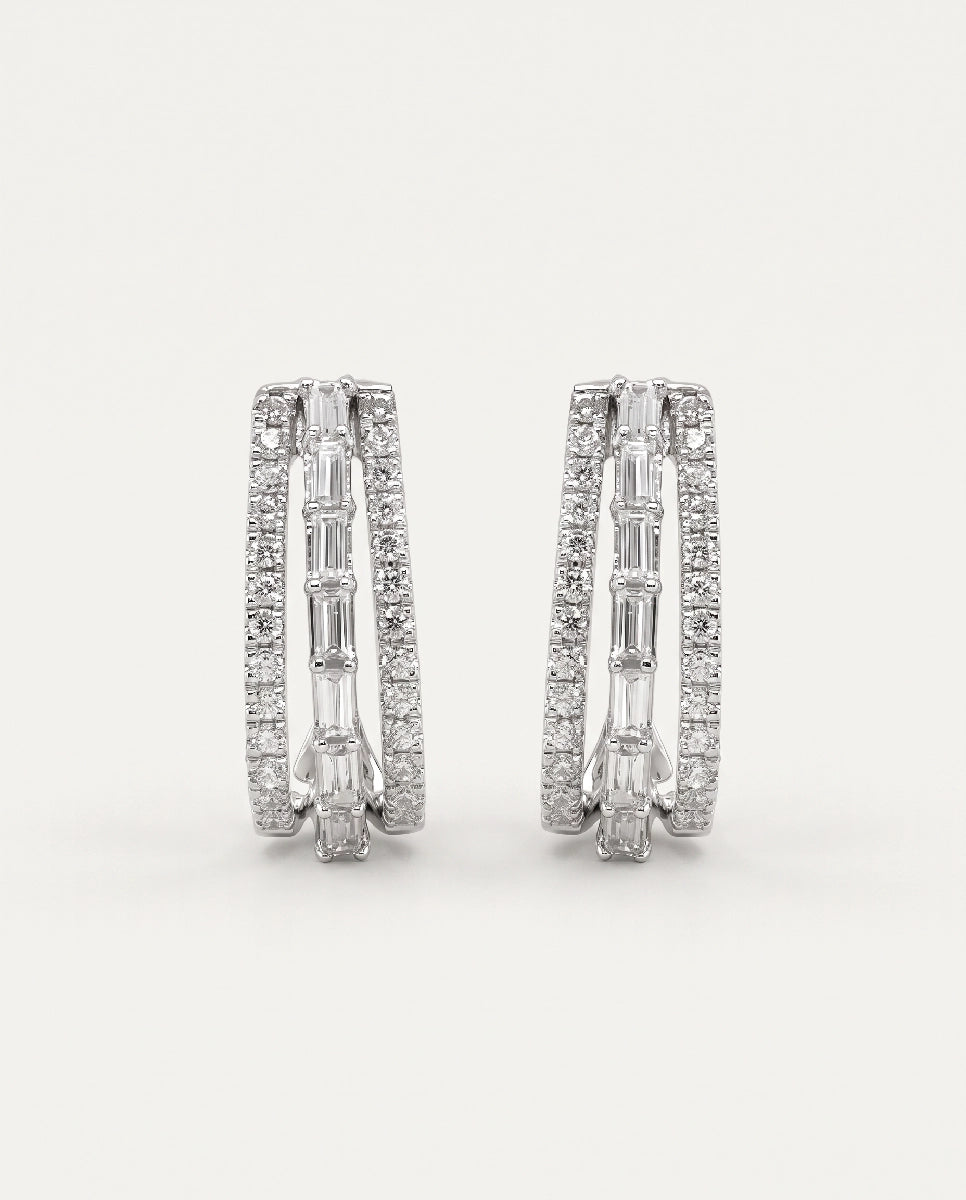 A pair of silver-toned huggie hoop earrings paved with small round and baguette-cut clear stones, shown against a plain light gray background.