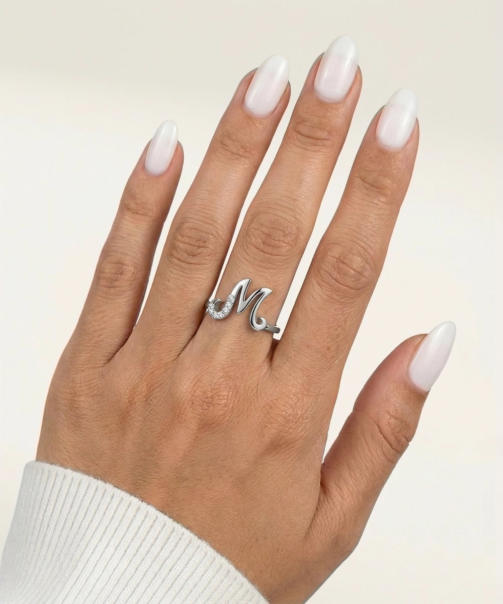 Hand wearing a silver ring with a letter 'M' on a beige background