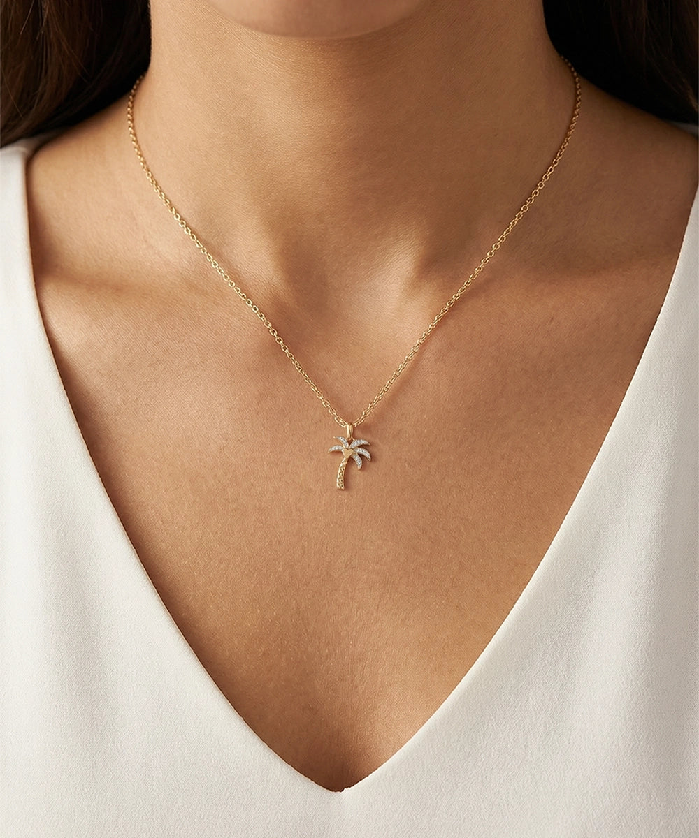 Gold necklace with a palm tree pendant on a person wearing a white top.