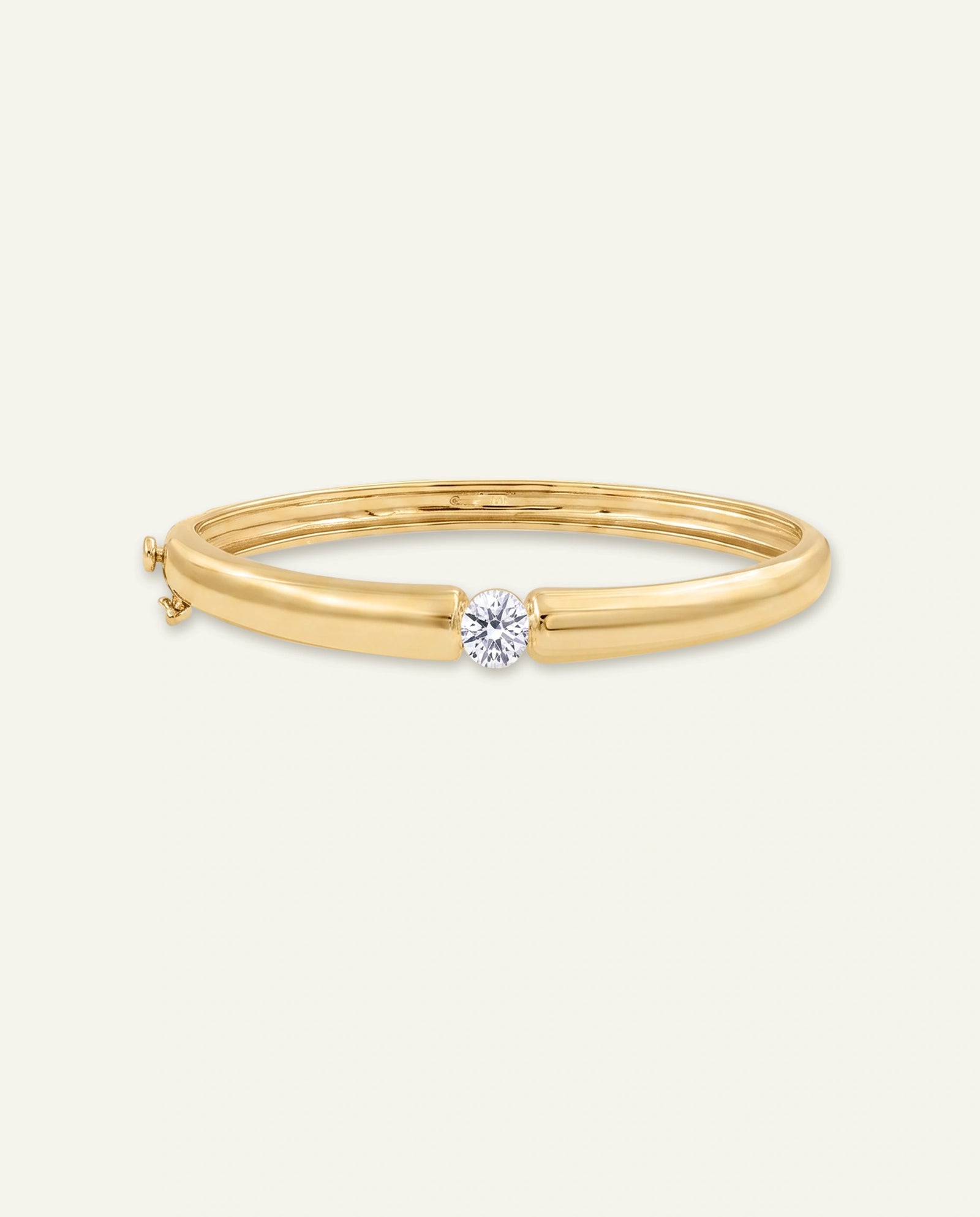 A polished yellow gold bangle bracelet with a single round-cut clear gemstone set in a bezel, shown on a light cream background.