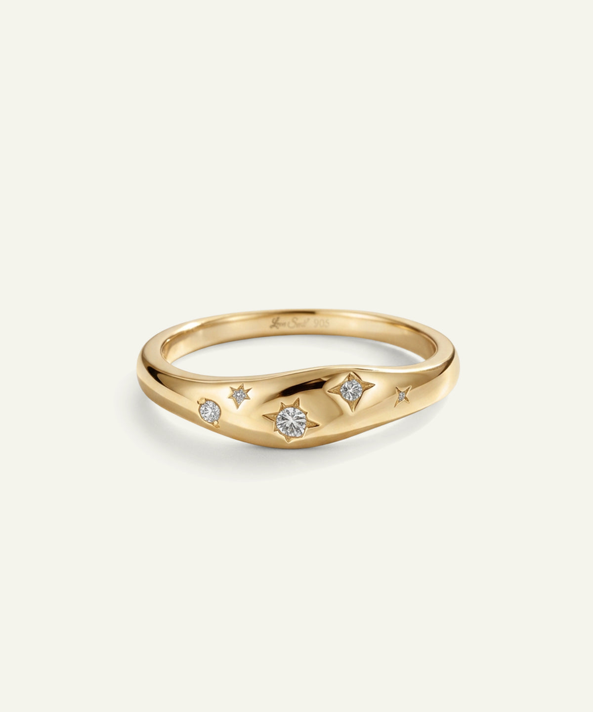 A gold ring with a wavy, shield-like shape featuring three small star motifs and a central round clear gemstone, set against a light off-white background.