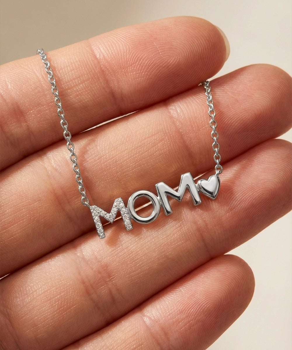 Silver 'MOM' necklace with a heart pendant held in a hand against a neutral background