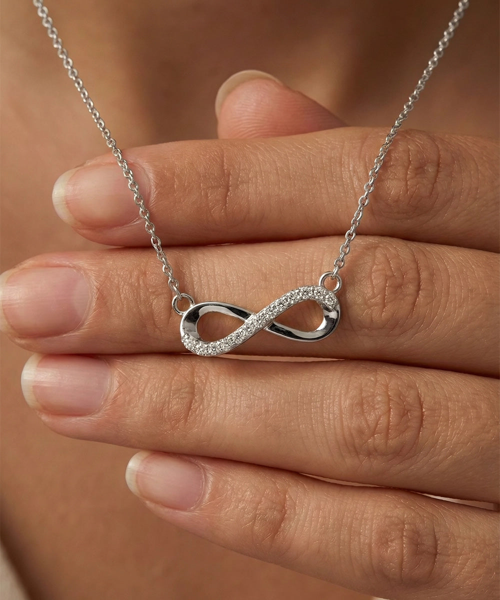 Silver infinity necklace held between fingers against a blurred background