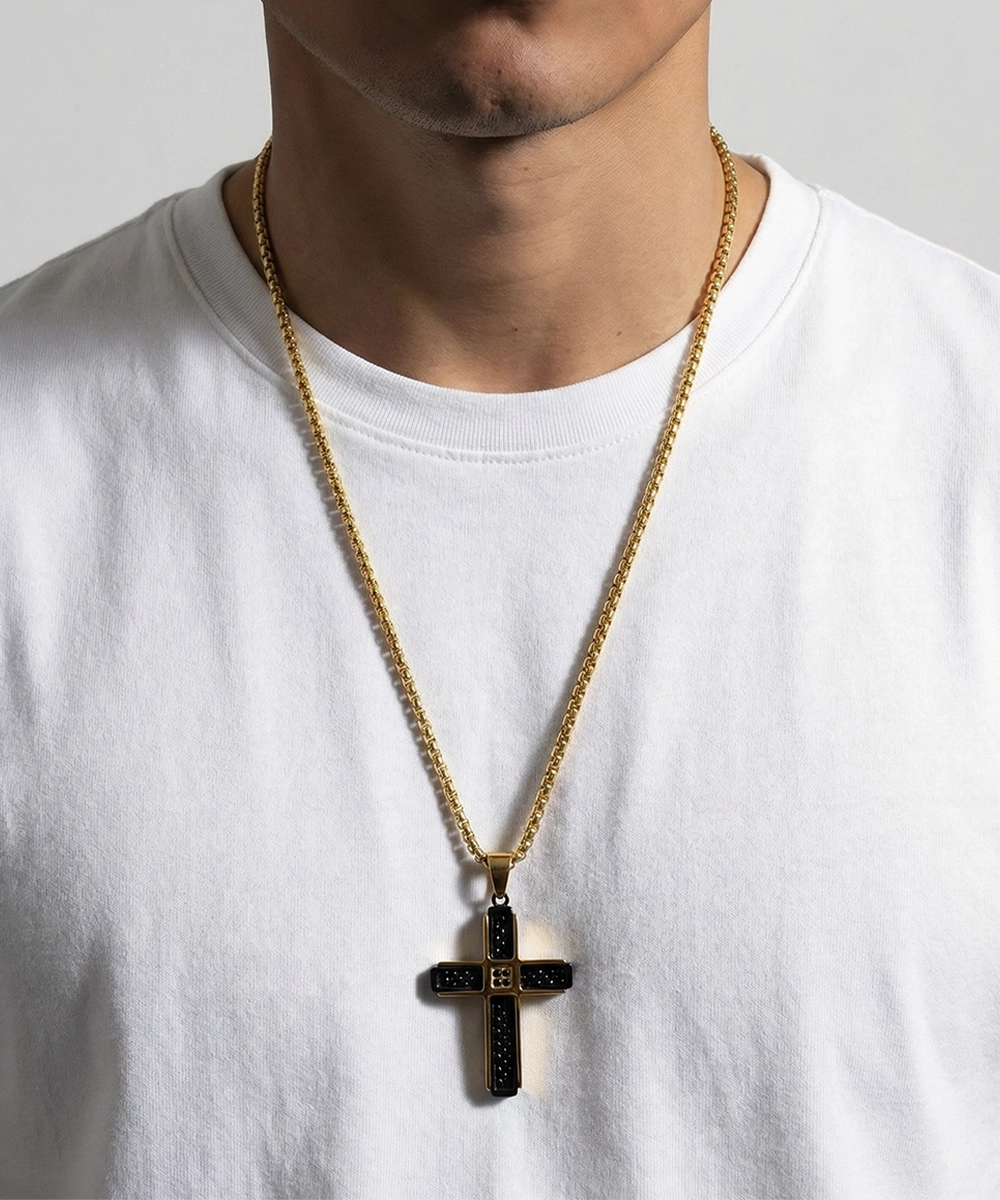 Person wearing a gold chain with a black cross pendant on a white shirt.