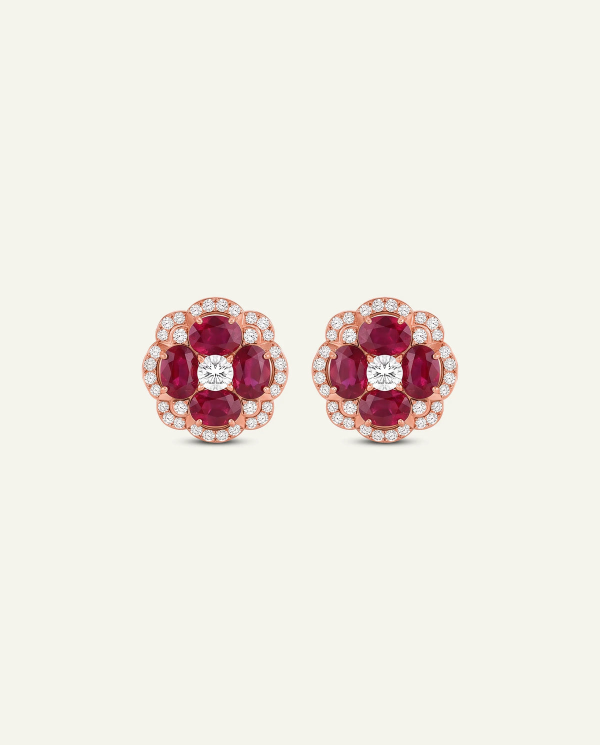A pair of rose gold stud earrings featuring oval-cut deep red rubies surrounded by small clear stones, with a round clear center stone, set against a solid off-white background.