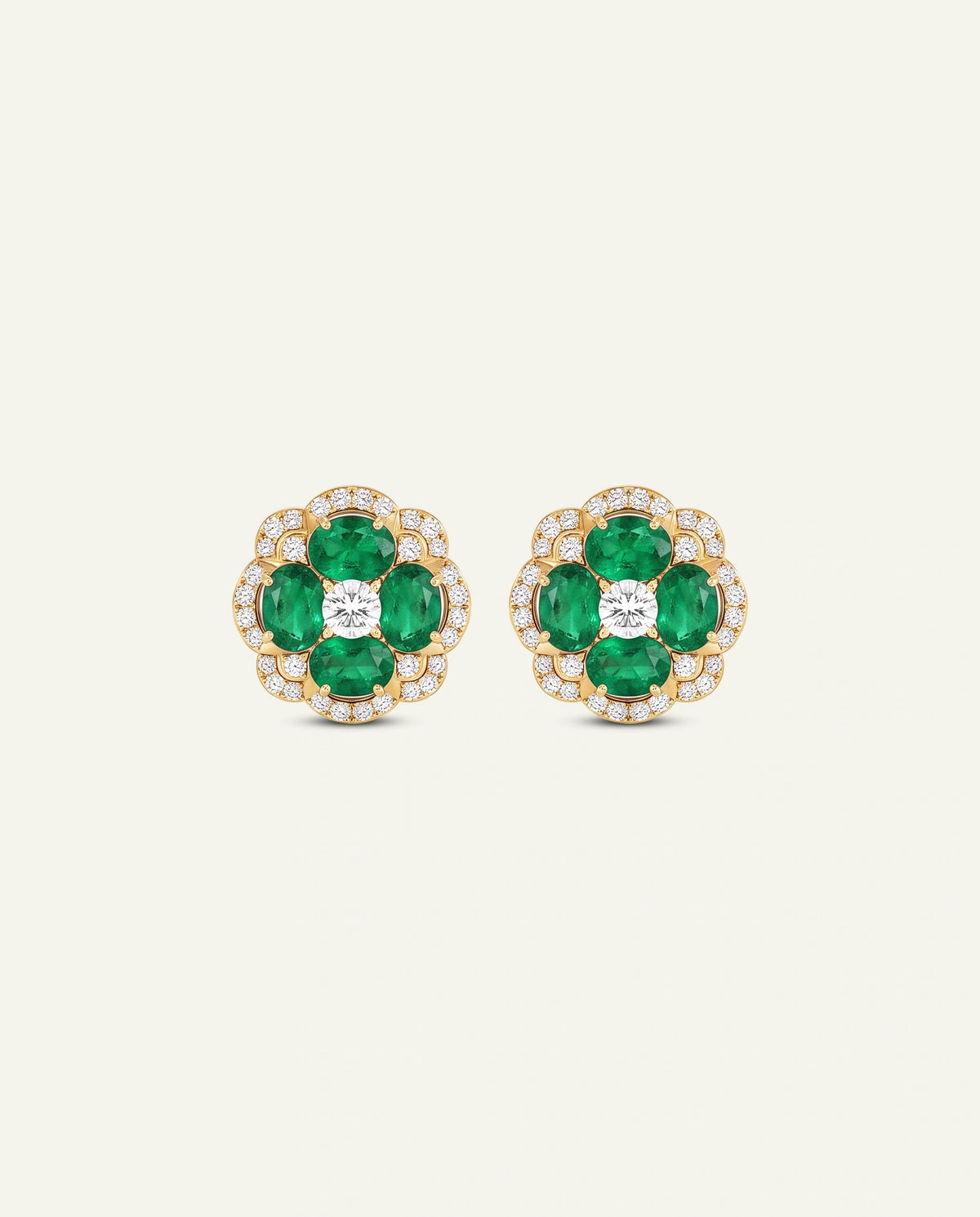 A pair of gold floral stud earrings featuring five green emerald-cut gemstones in each flower, with a central clear round-cut stone surrounded by small clear stones, set against a light cream background.