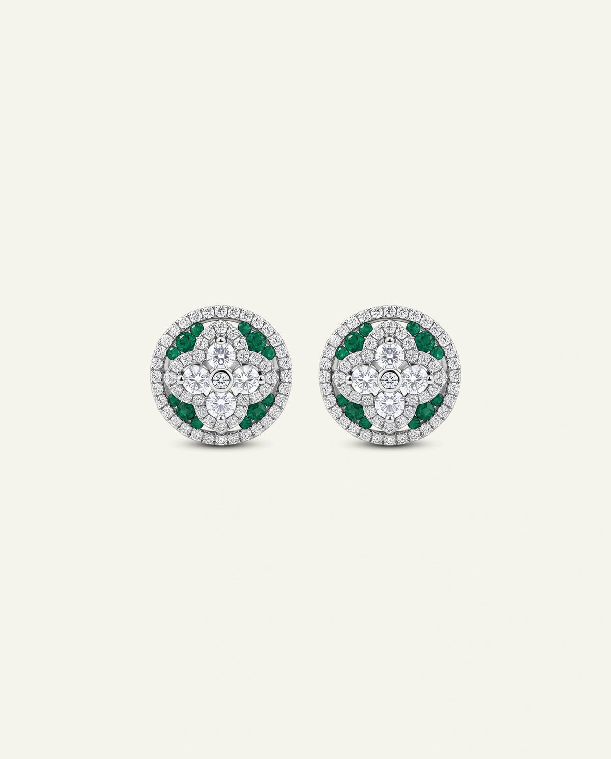 A pair of white gold round stud earrings featuring a center diamond cluster in a four-petal floral design, bordered by green emerald accents and an outer halo of pavÃ©-set diamonds, shown on a light off-white background.