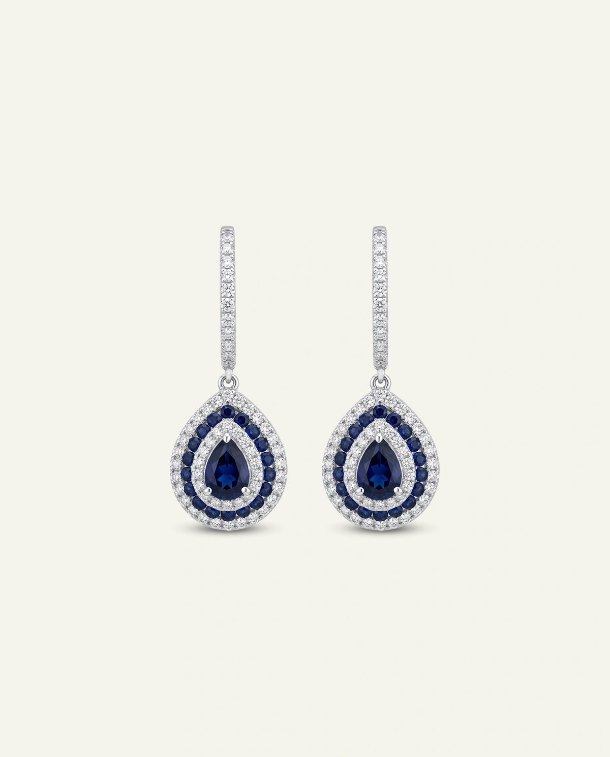 A pair of teardrop-shaped drop earrings in silver-tone metal, featuring a central deep blue sapphire surrounded by a halo of small clear stones, with a vertical bar of clear stones above, set against a light cream background.