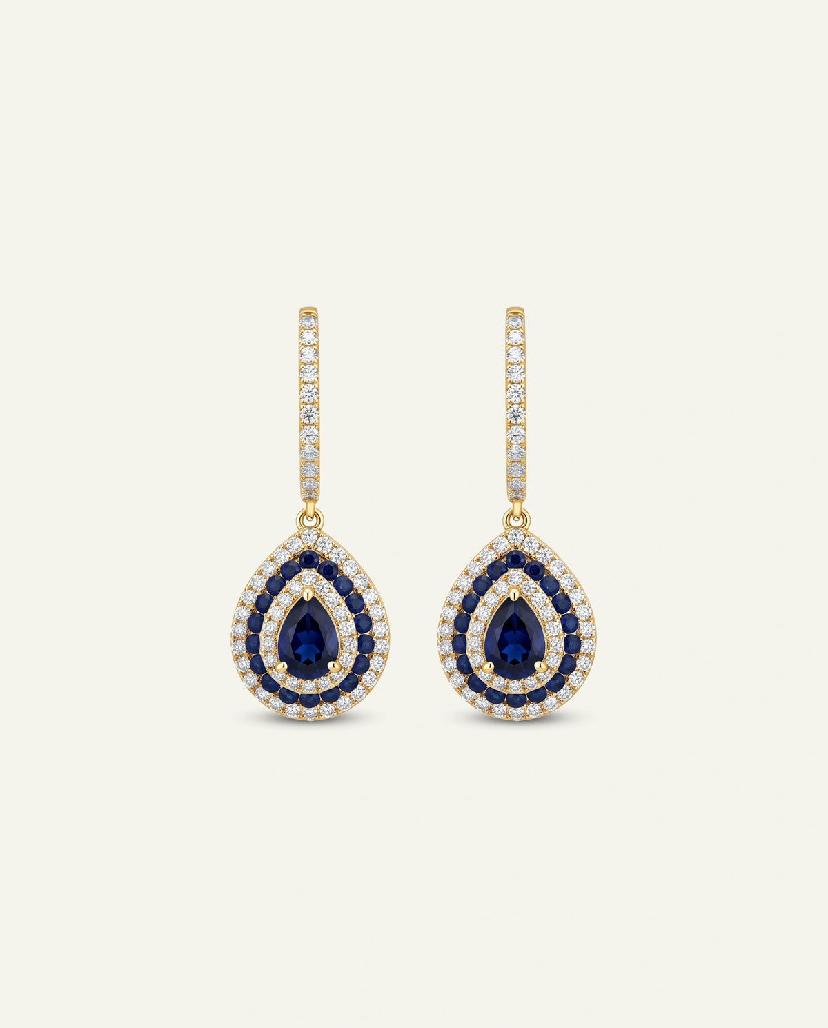 A pair of gold drop earrings featuring pear-shaped blue sapphires surrounded by double halos of clear stones, with a vertical bar of small clear stones above, set against a light cream background.