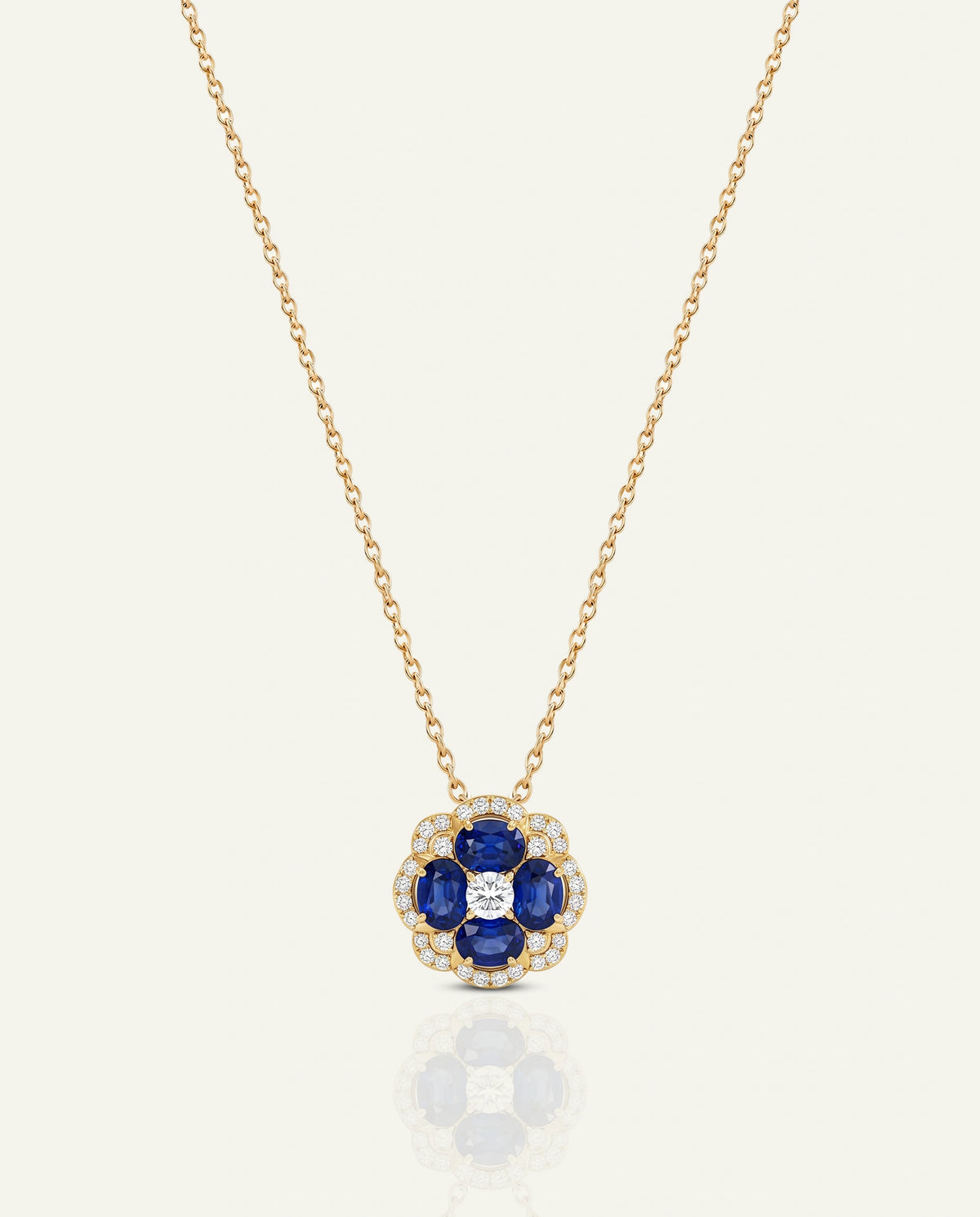 A gold chain necklace with a flower-shaped pendant featuring blue sapphires and a central clear diamond, set against a light cream background with a soft reflection below.