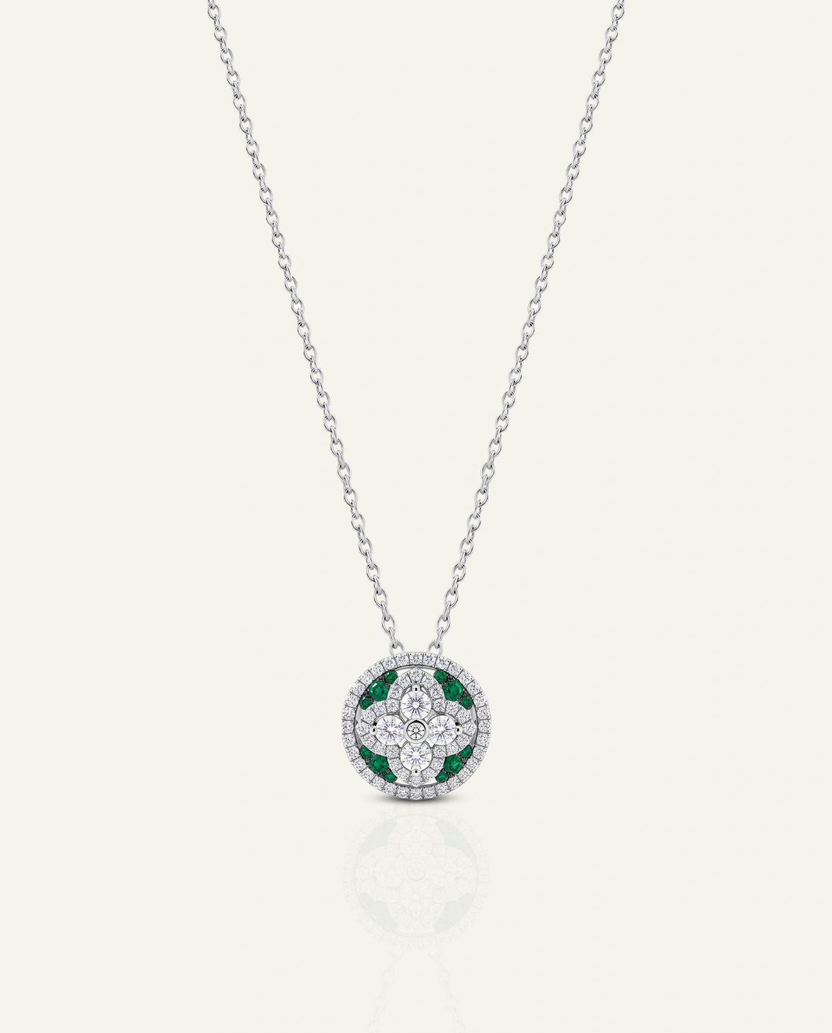 A delicate white gold chain necklace featuring a circular pendant with a center four-point floral cross motif set with clear diamonds and green emerald accents, enclosed by a diamond halo, shown on a light cream background.