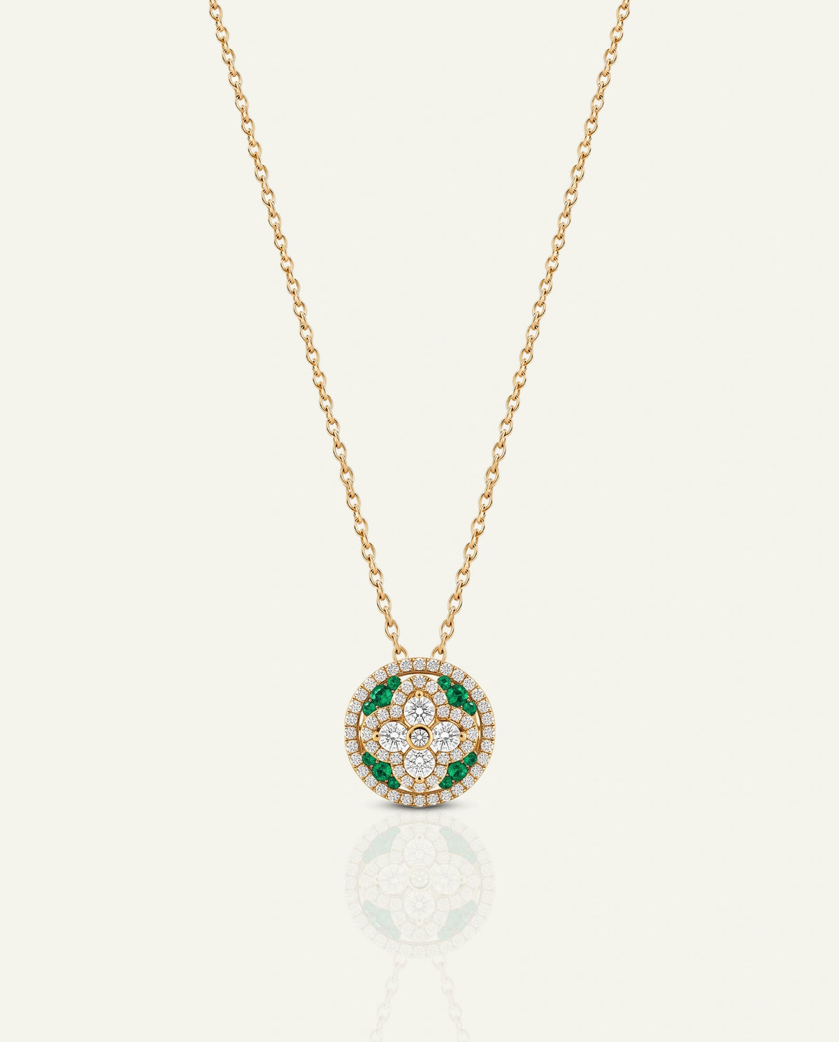 A delicate yellow gold chain necklace featuring a circular pendant with a center four-point floral cross motif set with clear diamonds and green emerald accents, enclosed by a diamond halo, shown on a light cream background.