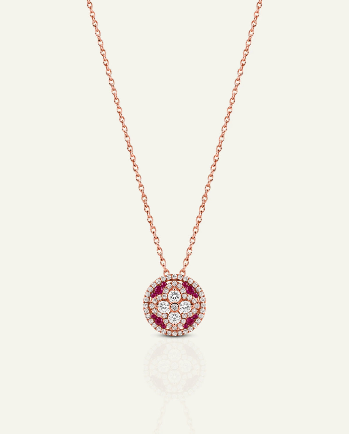 A rose gold necklace with a circular pendant featuring a floral pattern of red and clear gemstones, displayed on a light cream background with a soft reflection below.