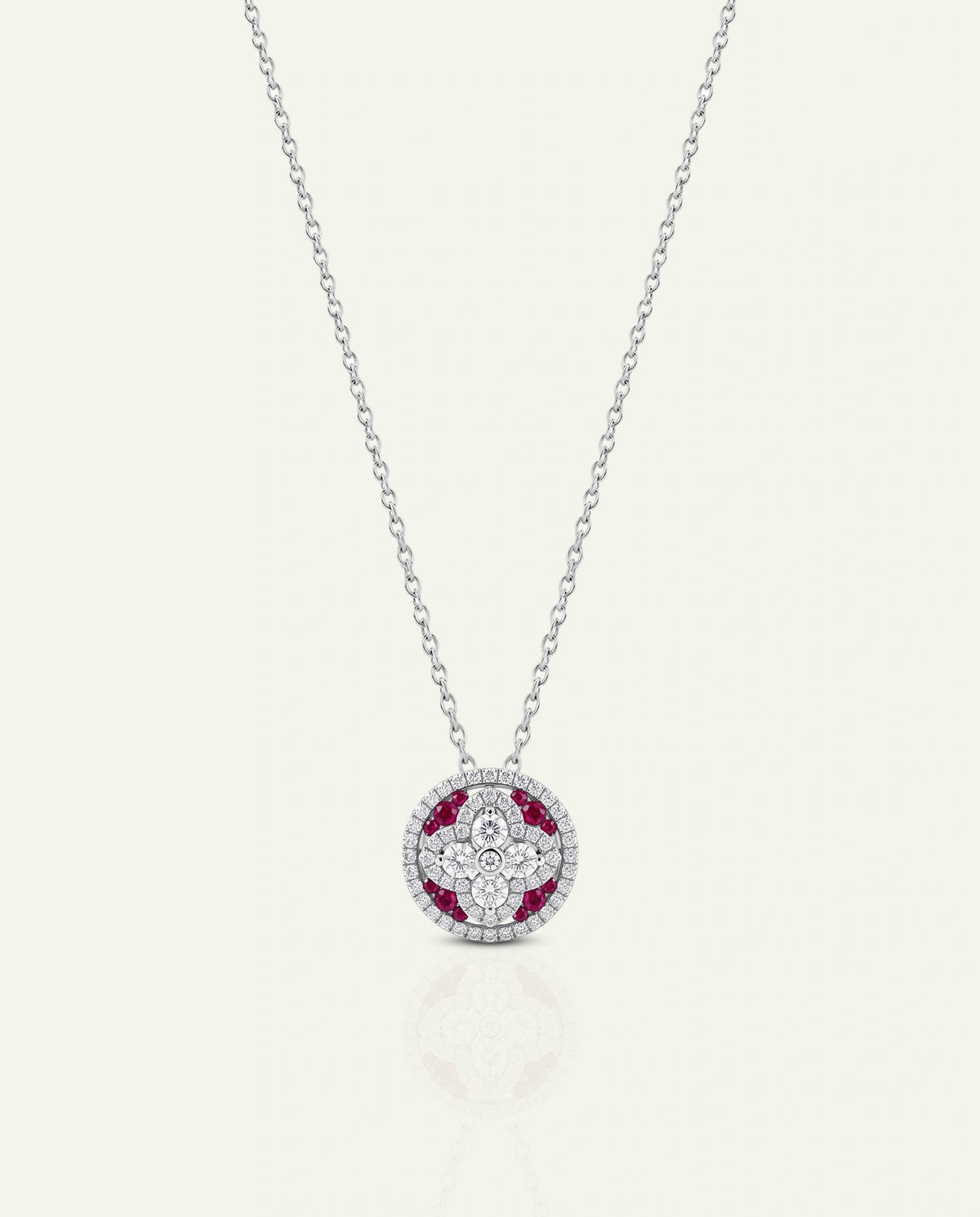 A delicate silver chain necklace with a circular pendant featuring a white floral pattern surrounded by red gemstones, set against a light cream background.