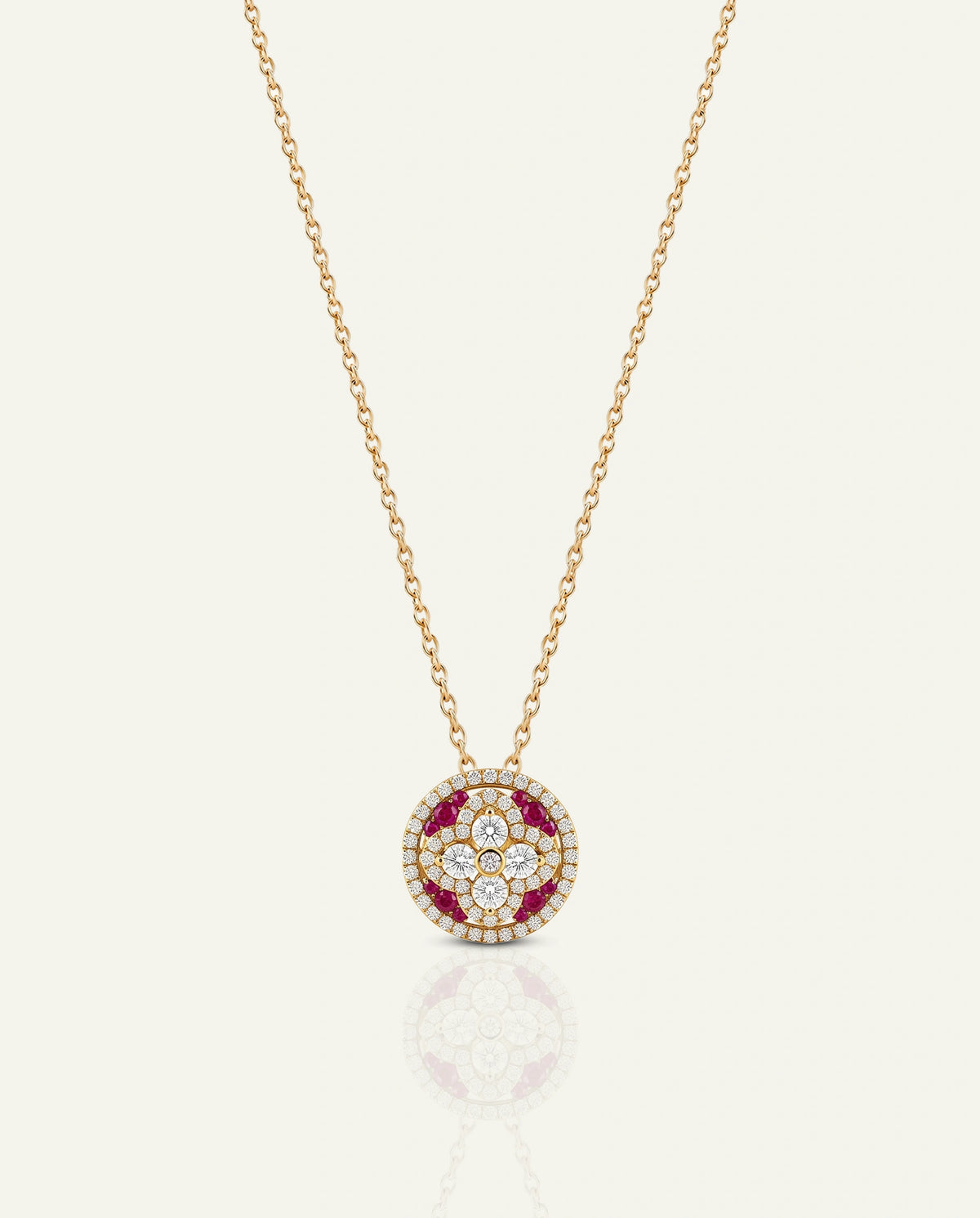 A gold chain necklace with a circular pendant featuring a floral pattern of white diamonds and a red gemstone border, displayed on a light cream background with a soft reflection.