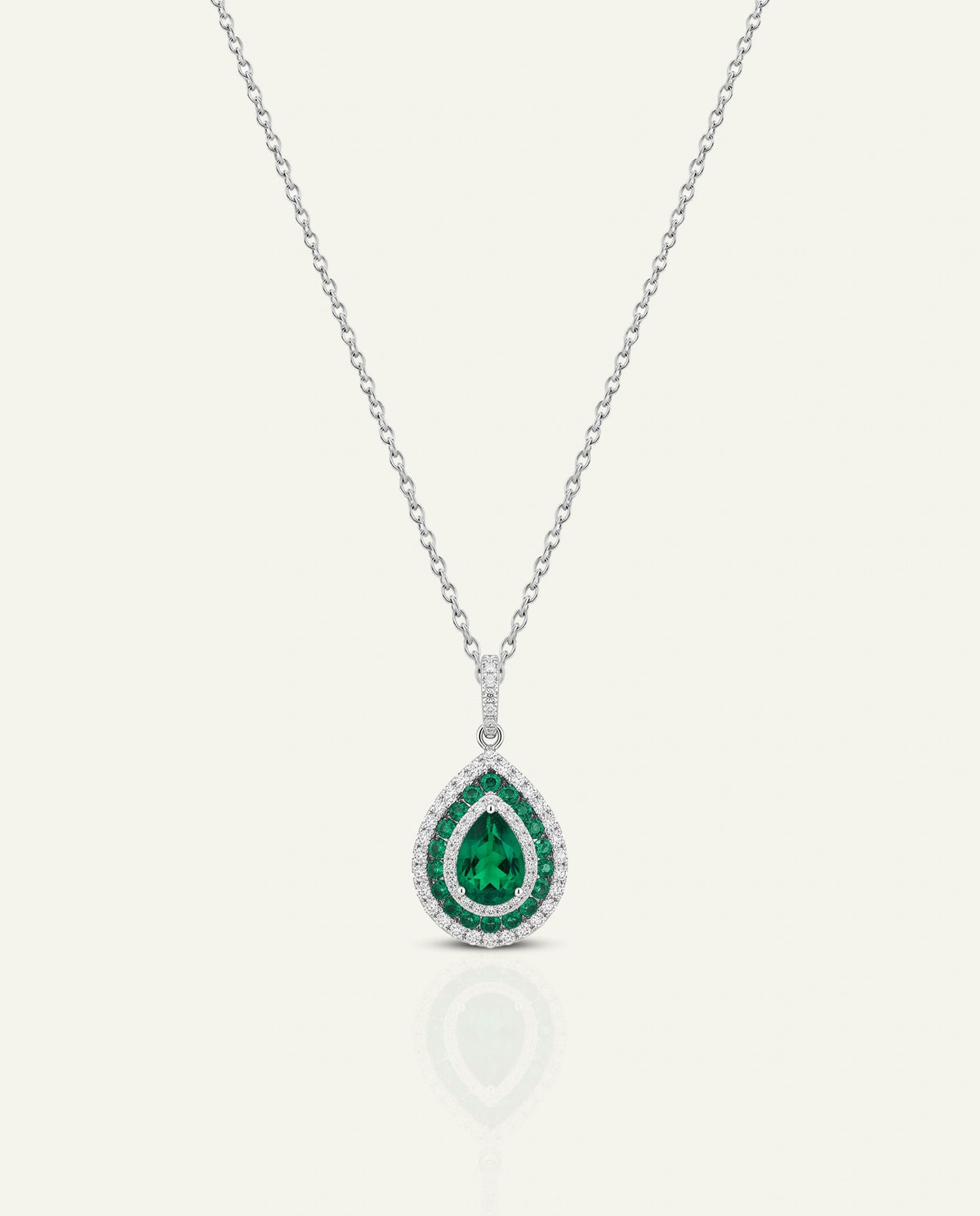 A silver-tone pendant necklace featuring a teardrop-shaped green emerald center stone surrounded by a double halo of small clear diamonds, suspended from a delicate chain on a light cream background.