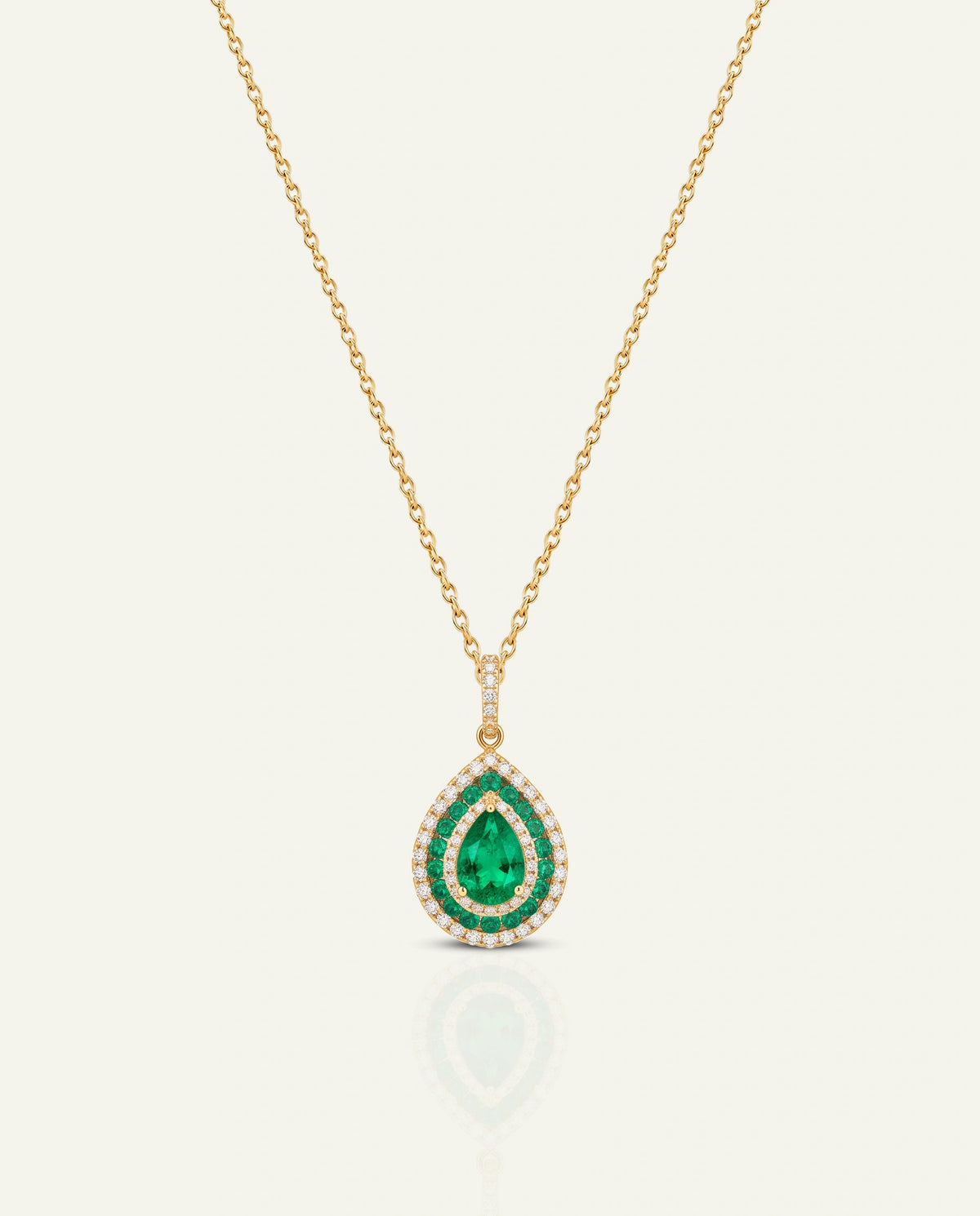 A gold chain necklace with a teardrop-shaped pendant featuring a central green emerald surrounded by a halo of small clear stones, set against a light cream background.
