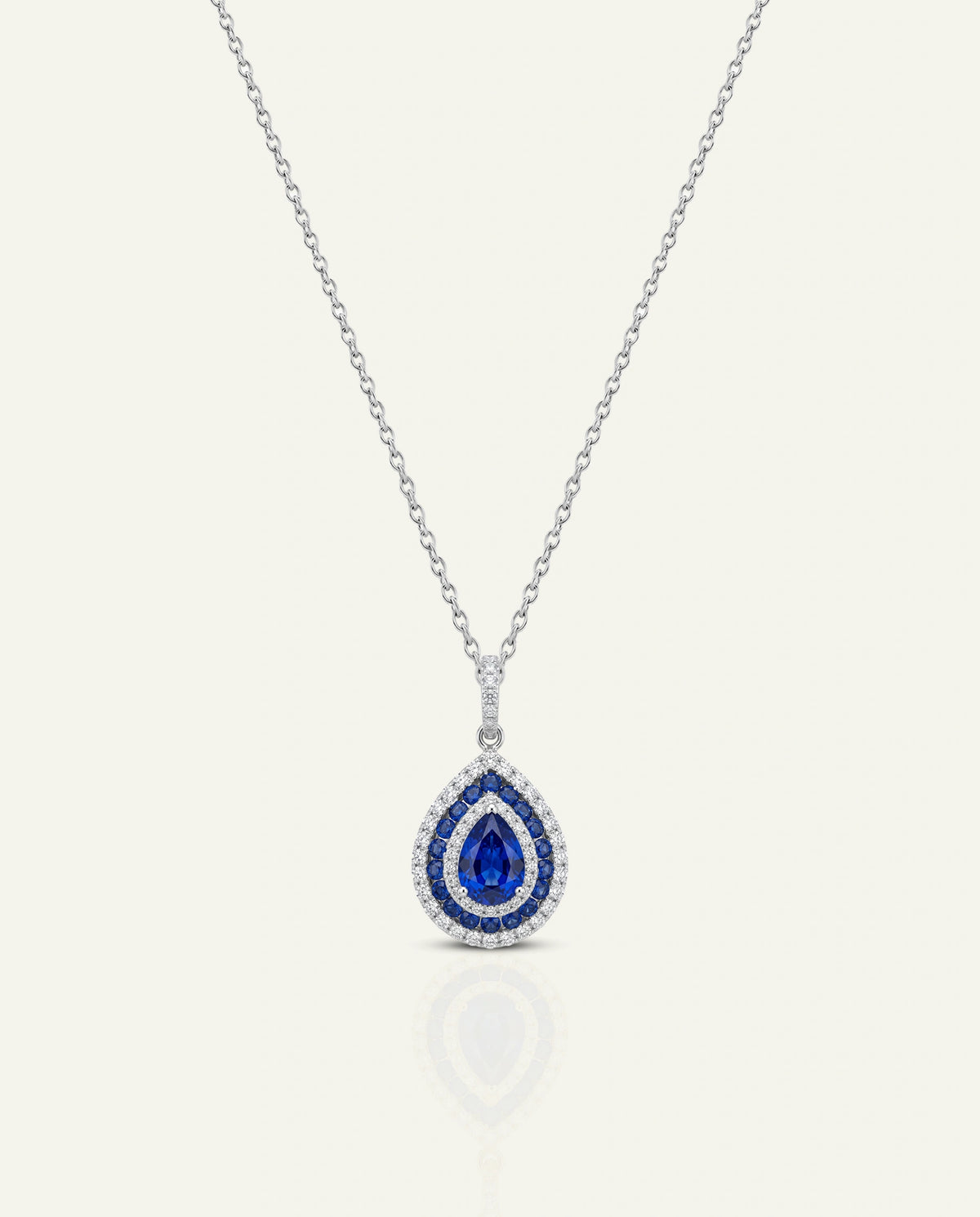 A silver-tone teardrop pendant necklace featuring a central deep blue sapphire surrounded by a halo of small clear stones, displayed on a light cream background with a soft reflection below.