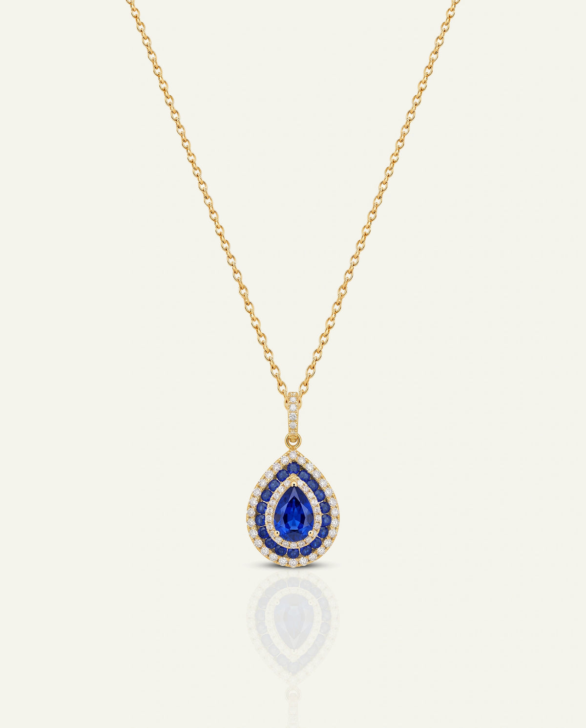 A gold chain necklace with a teardrop-shaped pendant featuring a central deep blue gemstone surrounded by small clear stones, shown on a light cream background with a soft reflection below.