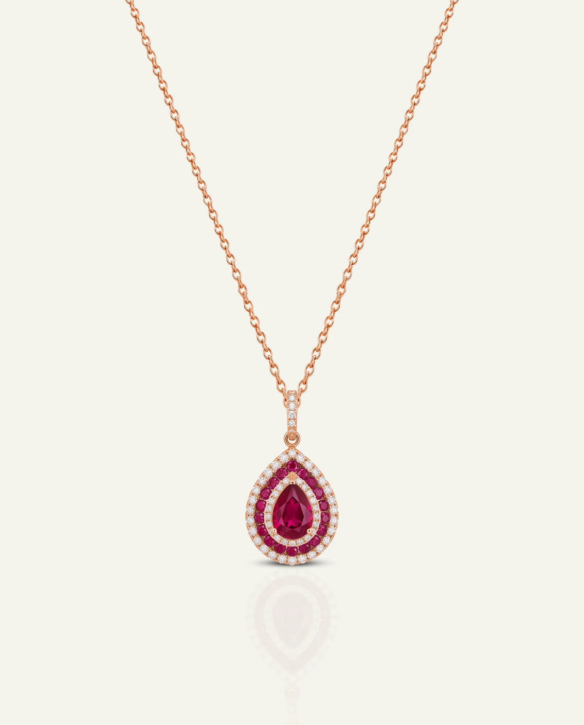 A rose gold teardrop pendant necklace featuring a central deep pinkish-red teardrop gemstone surrounded by a halo of small clear stones, suspended from a delicate rose gold chain on a light cream background.