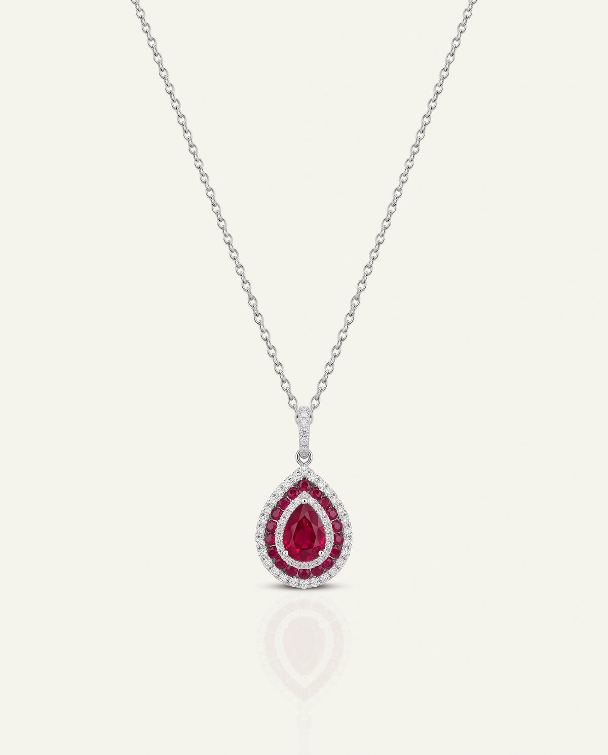A silver-tone teardrop pendant necklace featuring a central deep red pear-cut gemstone surrounded by a halo of small clear stones, suspended on a delicate chain against a light cream background.