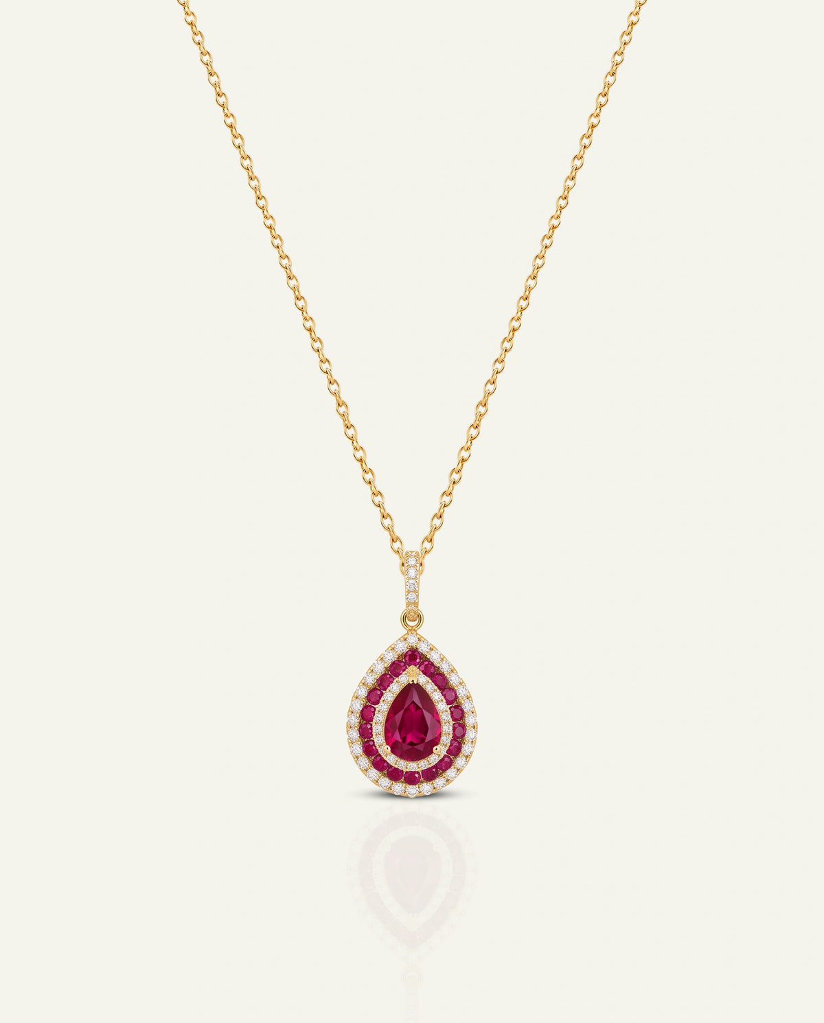A gold pendant necklace featuring a teardrop-shaped ruby pendant surrounded by a halo of small white stones, suspended from a delicate gold chain on a light cream background.