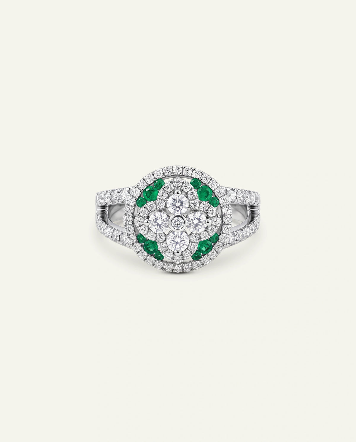 A silver-toned ring featuring a circular floral cluster with green emerald-cut stones and clear pavÃ©-set stones, centered on a light cream background.